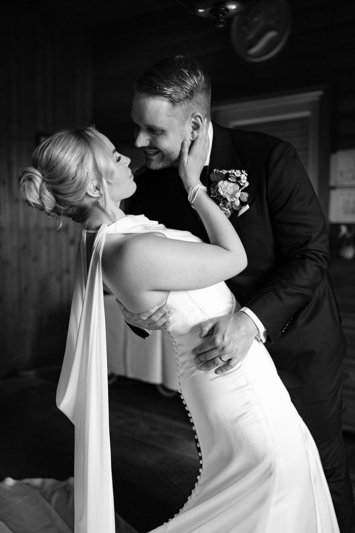 Black and white photo of a smiling groom in a suit embracing his bride in a white gown during their wedding day.