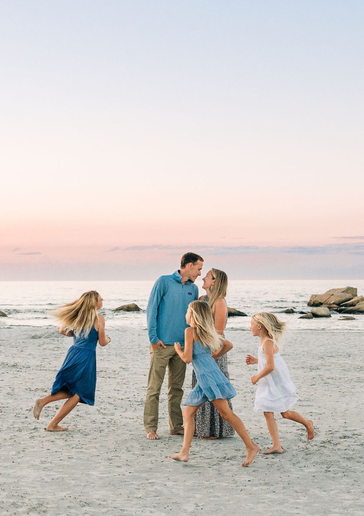 Family of five enjoying a romantic beach sunset, with children running around parents sharing a tender moment on sandy shore.