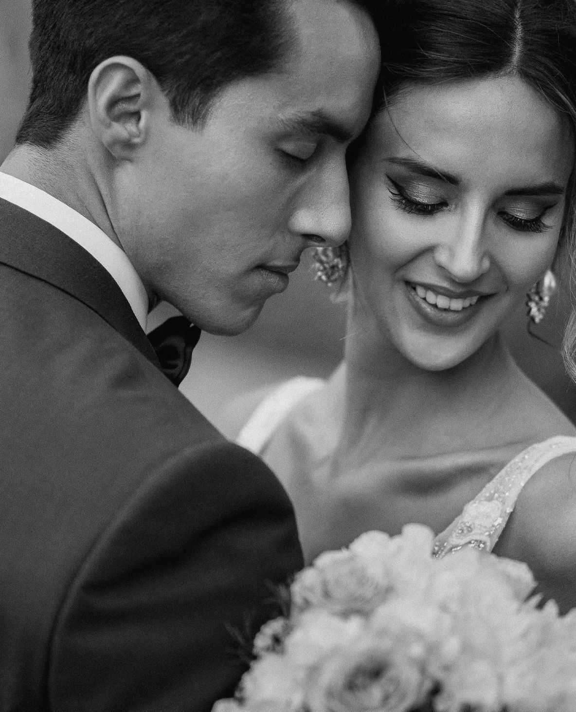 Intimate black and white portrait of groom nuzzling bride holding a white bouquet.