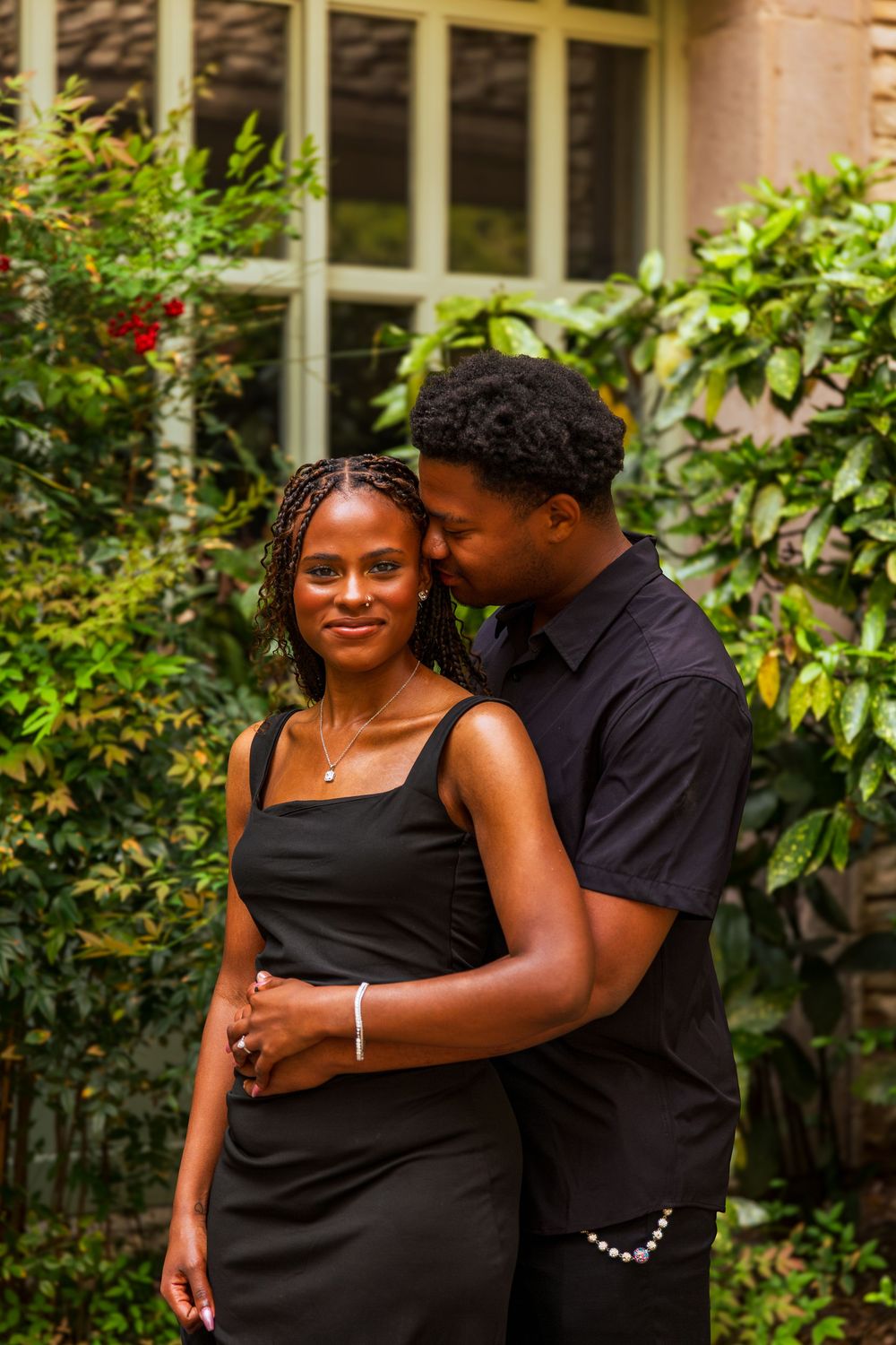 A couple in matching black outfits embraces tenderly in a lush garden, the man kissing the womans temple.