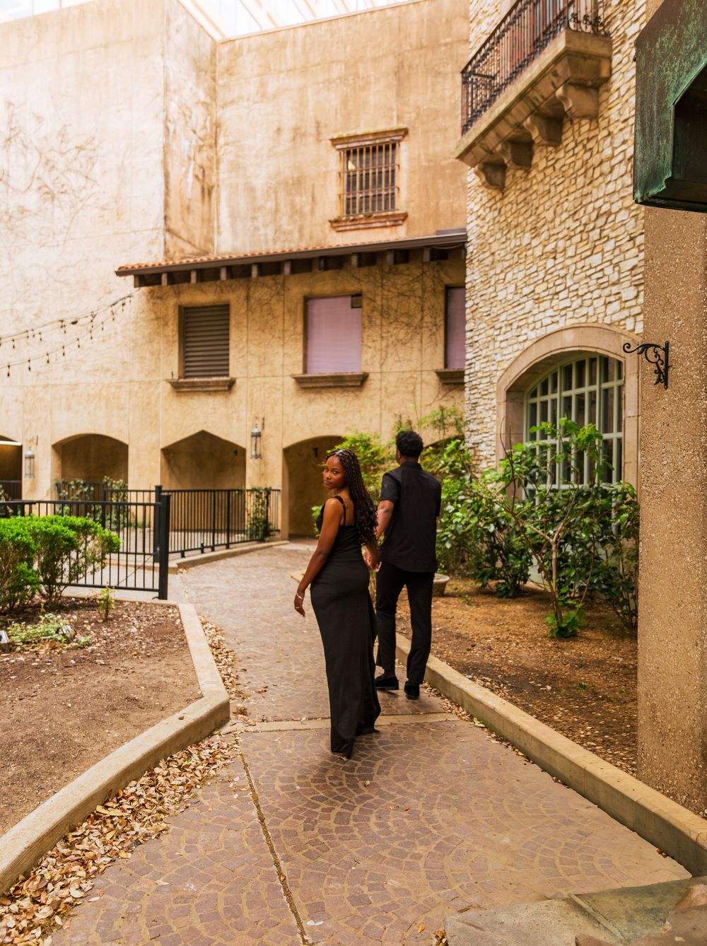Couple in formal black attire walking along a stone courtyard pathway surrounded by Mediterranean-style architecture.