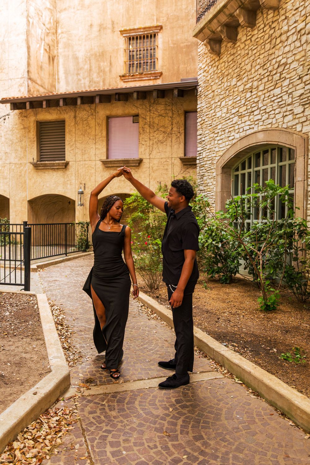 Couple dancing outdoors in black outfits, man spinning woman on cobblestone path near stone buildings.