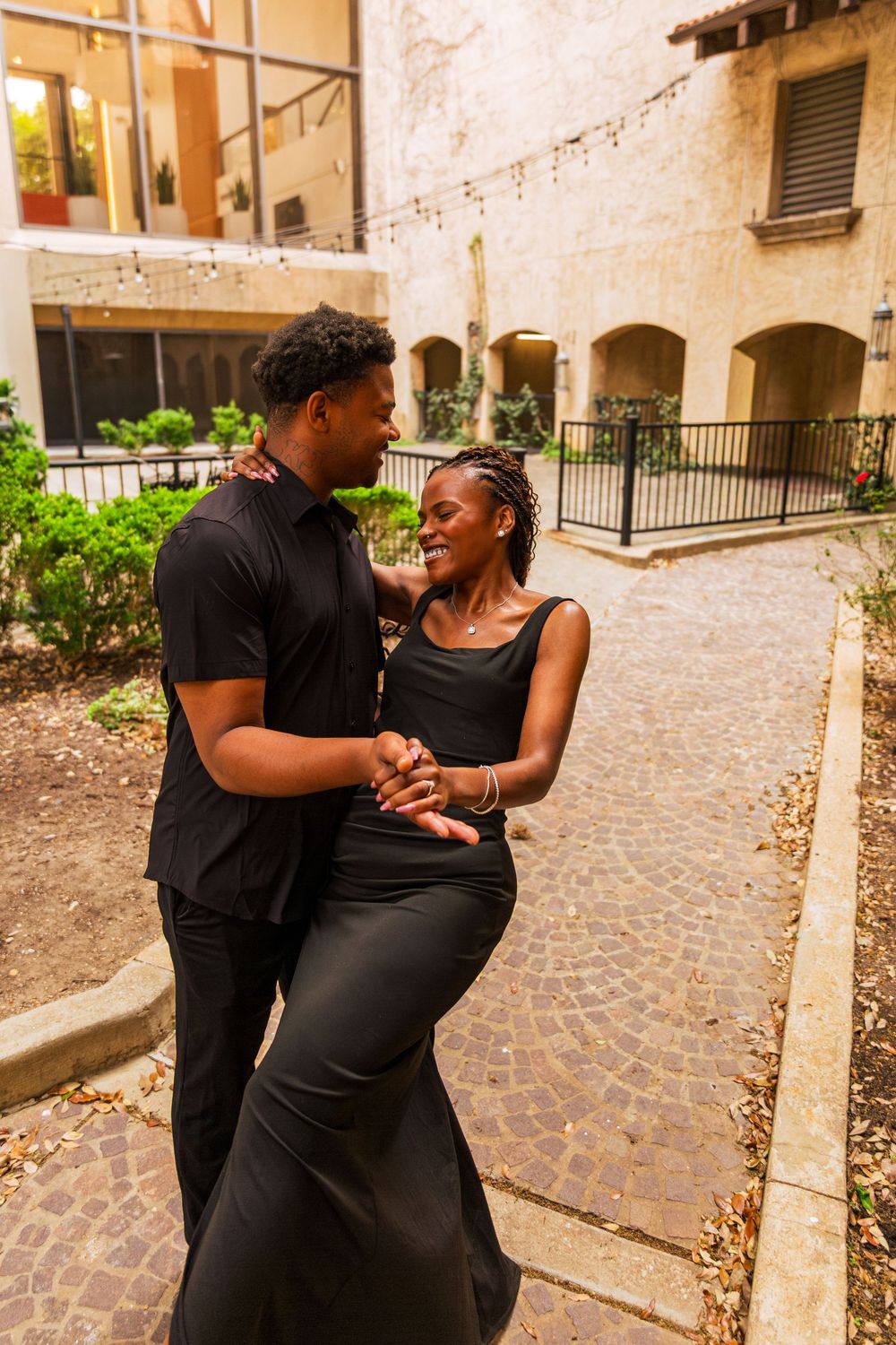 A joyful Black couple in matching black outfits share a romantic dance in a charming courtyard setting.