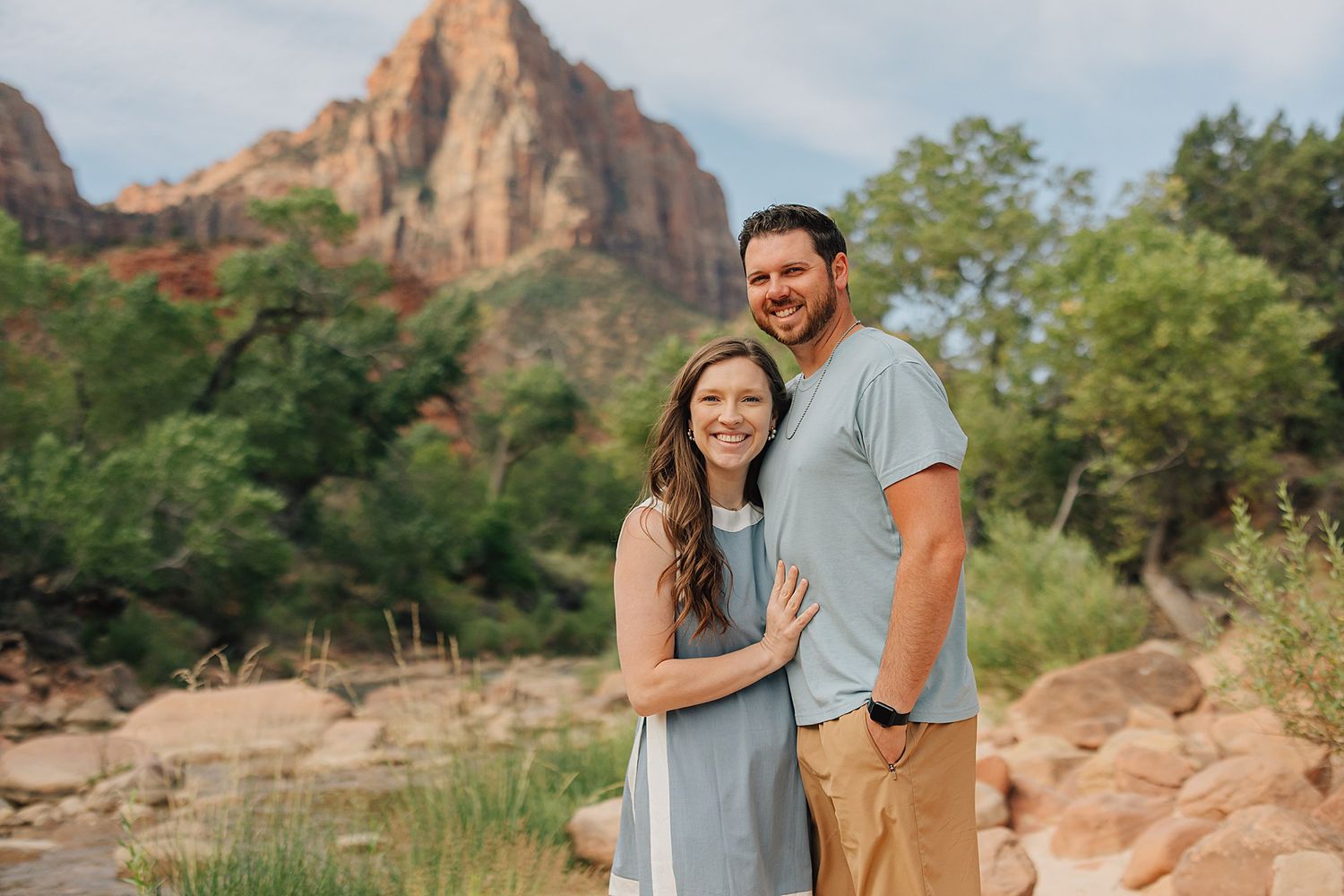 Couple smiling together in front of stunning Zion National Park mountain landscape with lush greenery.