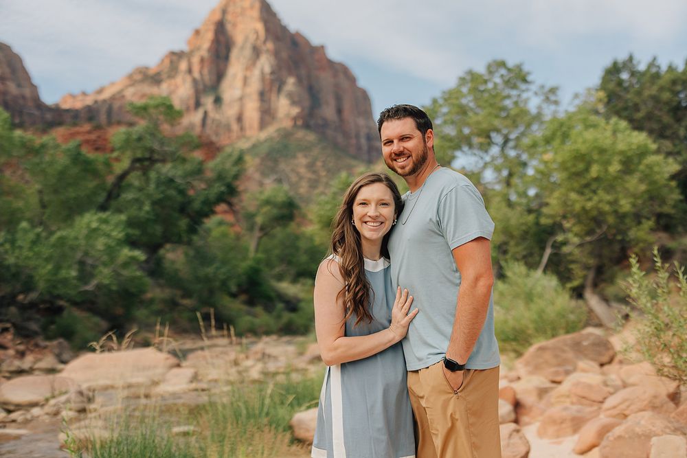 Couple smiling together in front of stunning Zion National Park mountain landscape with lush greenery.
