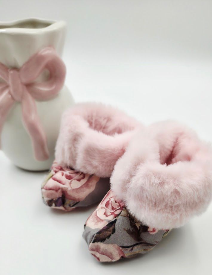 Pink floral baby booties with faux fur trim displayed next to a white ceramic vase with pink bow ribbon.