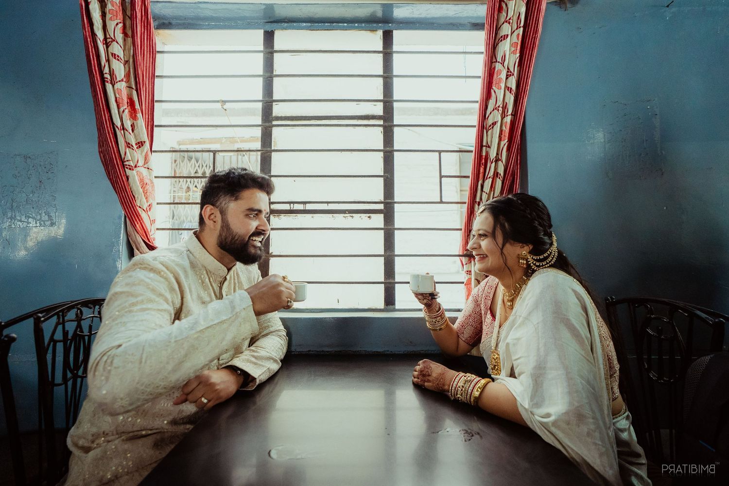 Indian couple in traditional attire sharing tea at a wooden table, smiling warmly in a blue room with red curtains.