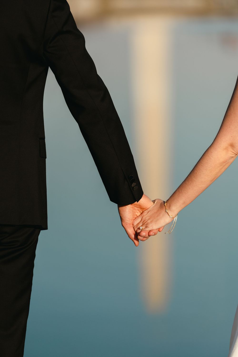 Couple holding hands in formal wedding attire near reflective water, showcasing a delicate bracelet and suit sleeve.