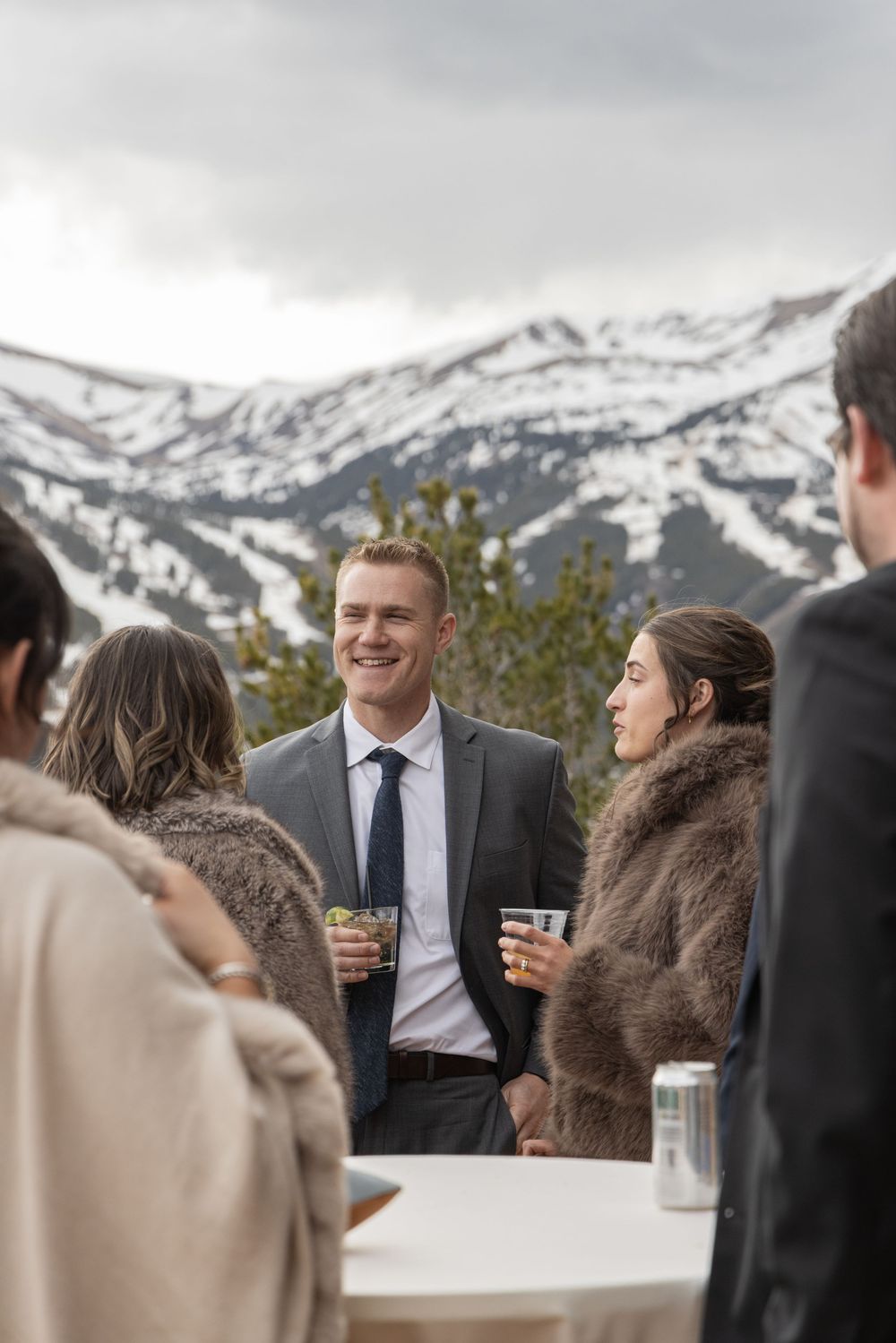 Guests in formal attire socializing outdoors at a mountain wedding reception with snow-capped peaks in the background.