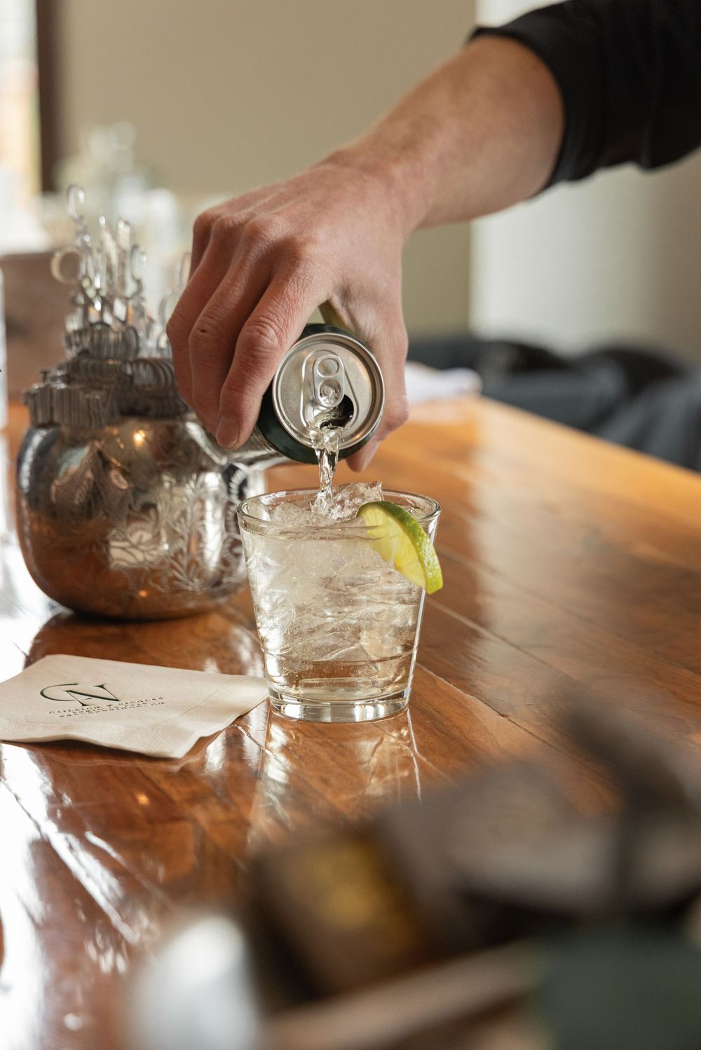 Bartender pouring tonic water from a can into a glass with lime wedge on a wooden bar.