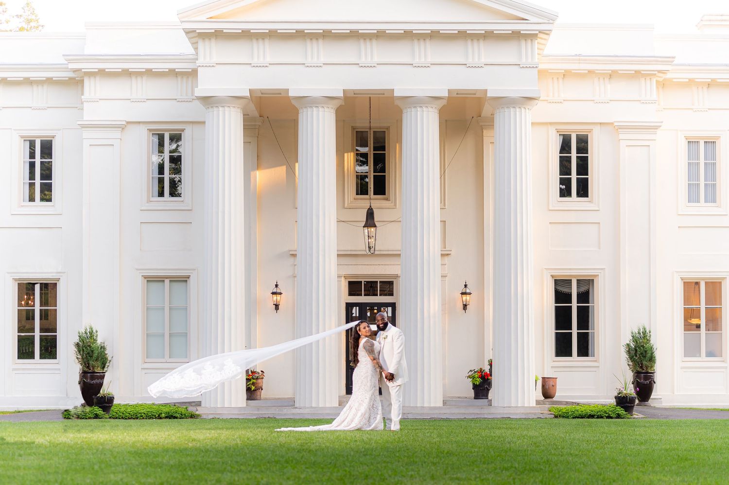 Bride and groom kiss in front of grand white columned mansion with flowing veil on lush green lawn.
