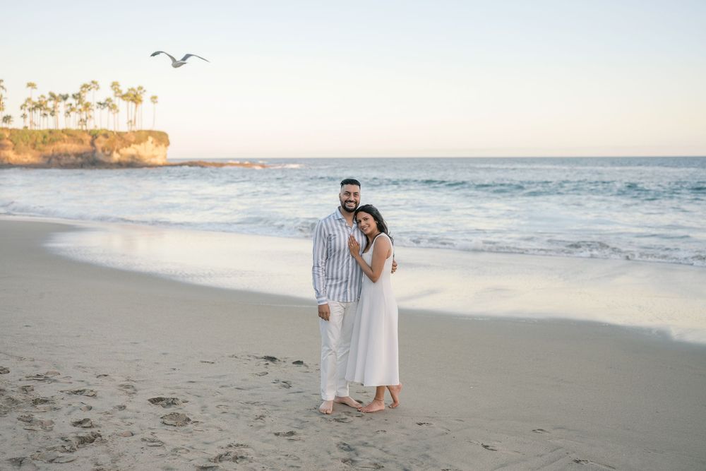 Couple in white outfits posing on a sandy beach at sunset with ocean waves and palm trees in background.