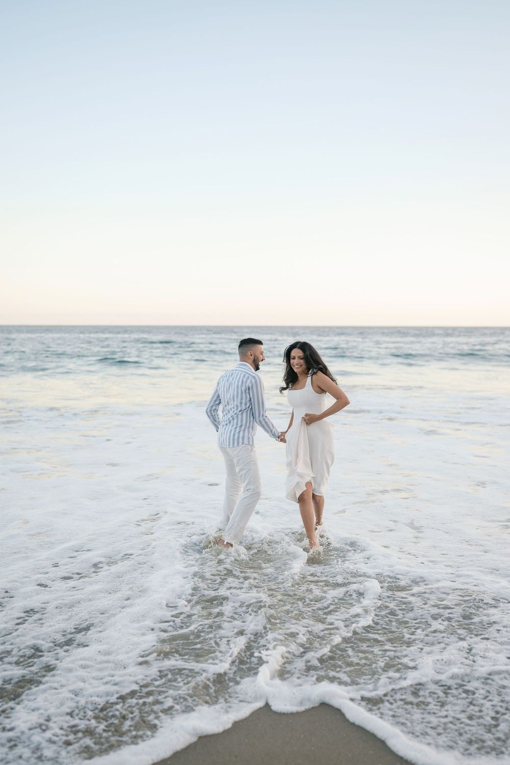 A couple holds hands and walks through ocean waves on a sandy beach at sunset, wearing elegant white outfits.