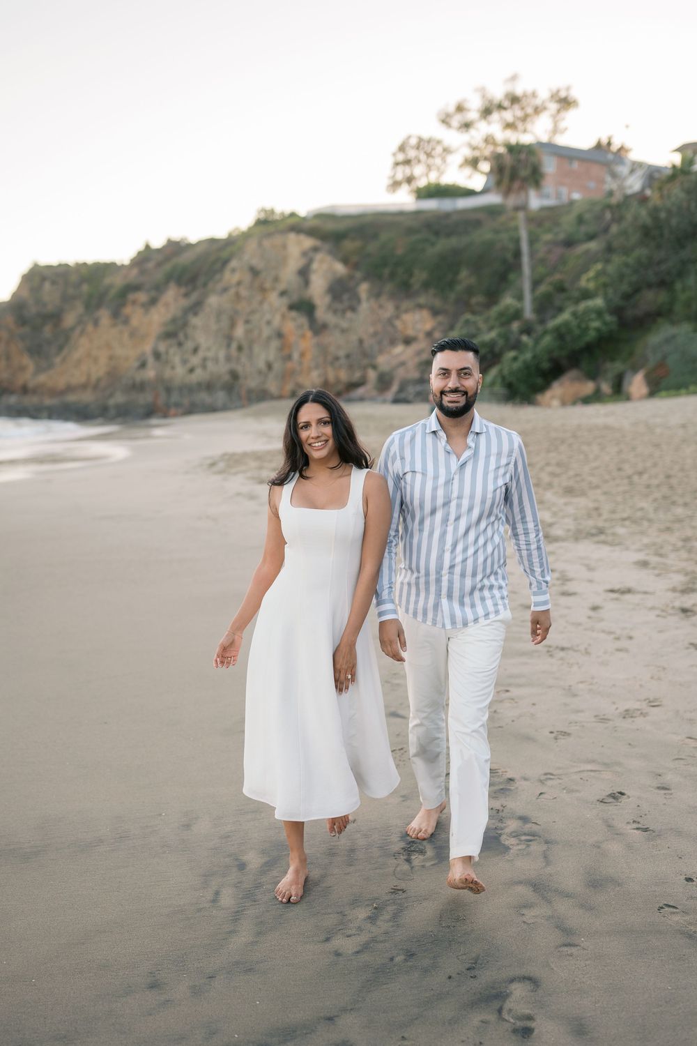 Couple walking barefoot on sandy beach, woman in white dress, man in striped shirt and white pants, cliffs in background.