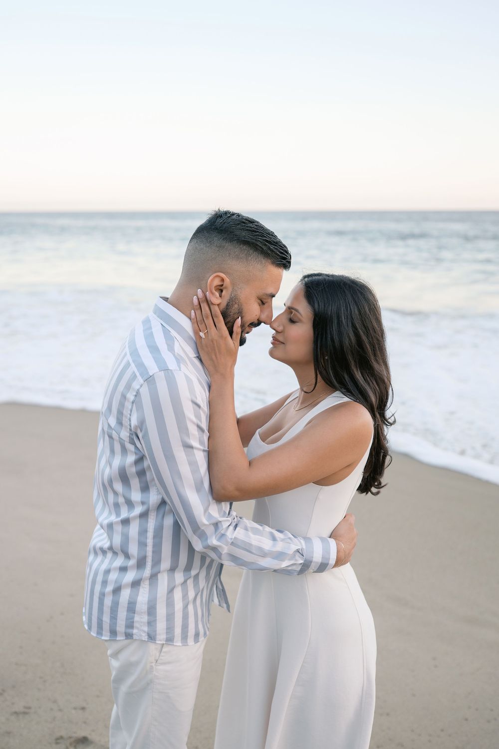 Romantic couple sharing an intimate moment on a beach at sunset, dressed in white and striped outfits.