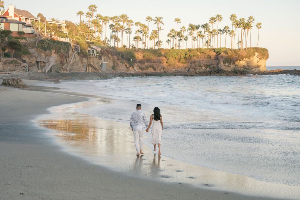 Couple holding hands walking barefoot on a sandy beach at sunset with palm trees and cliffs in the background.