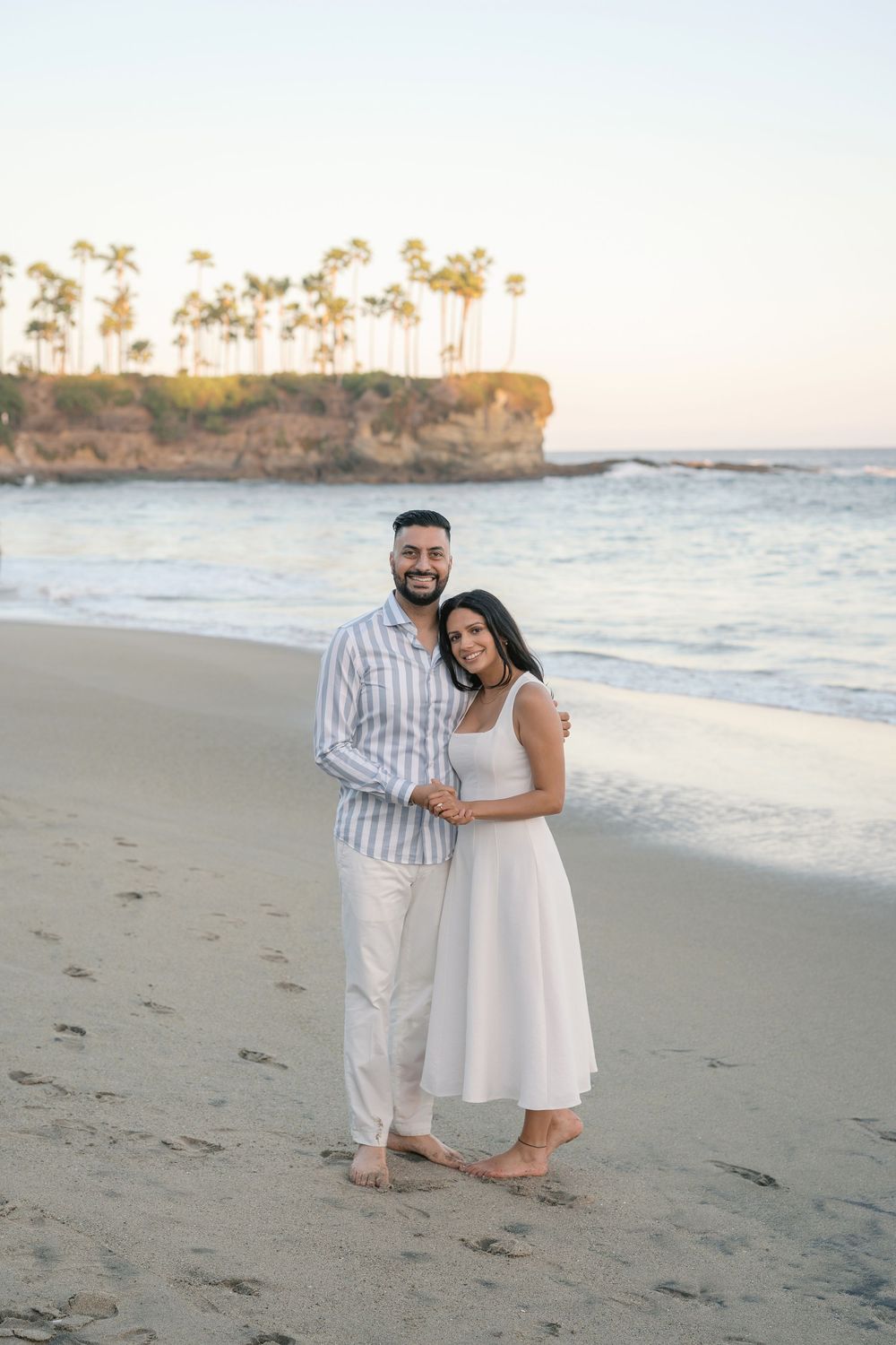 Couple embracing on a sandy beach at sunset with palm trees and ocean waves in the background.