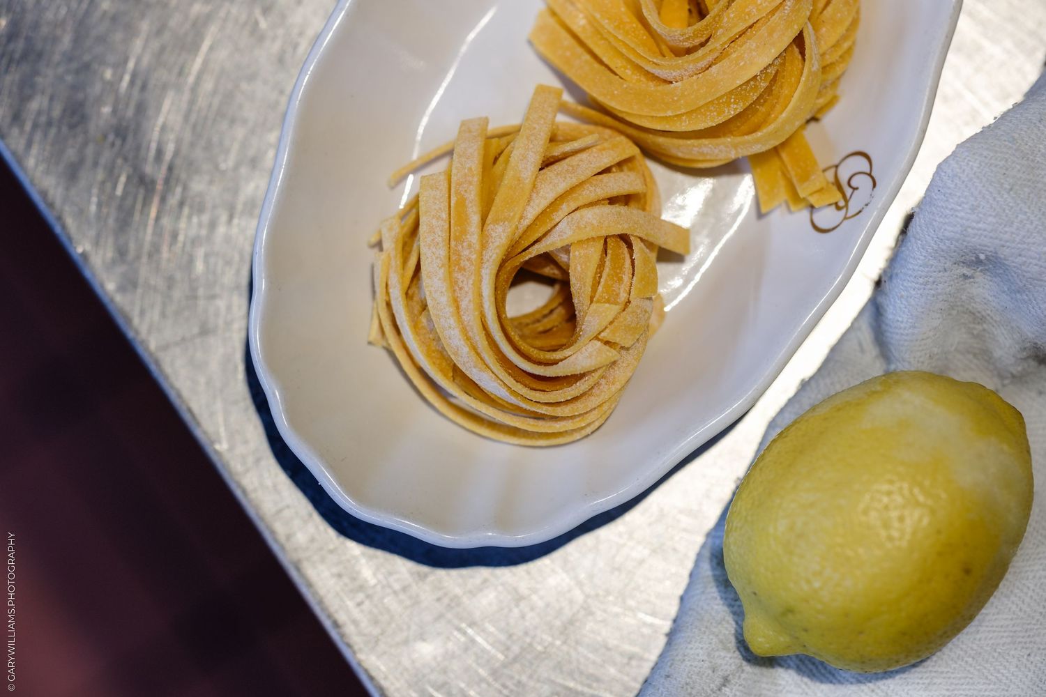 Two nests of fresh fettuccine pasta on a white oval plate beside a bright yellow lemon on a gray surface.