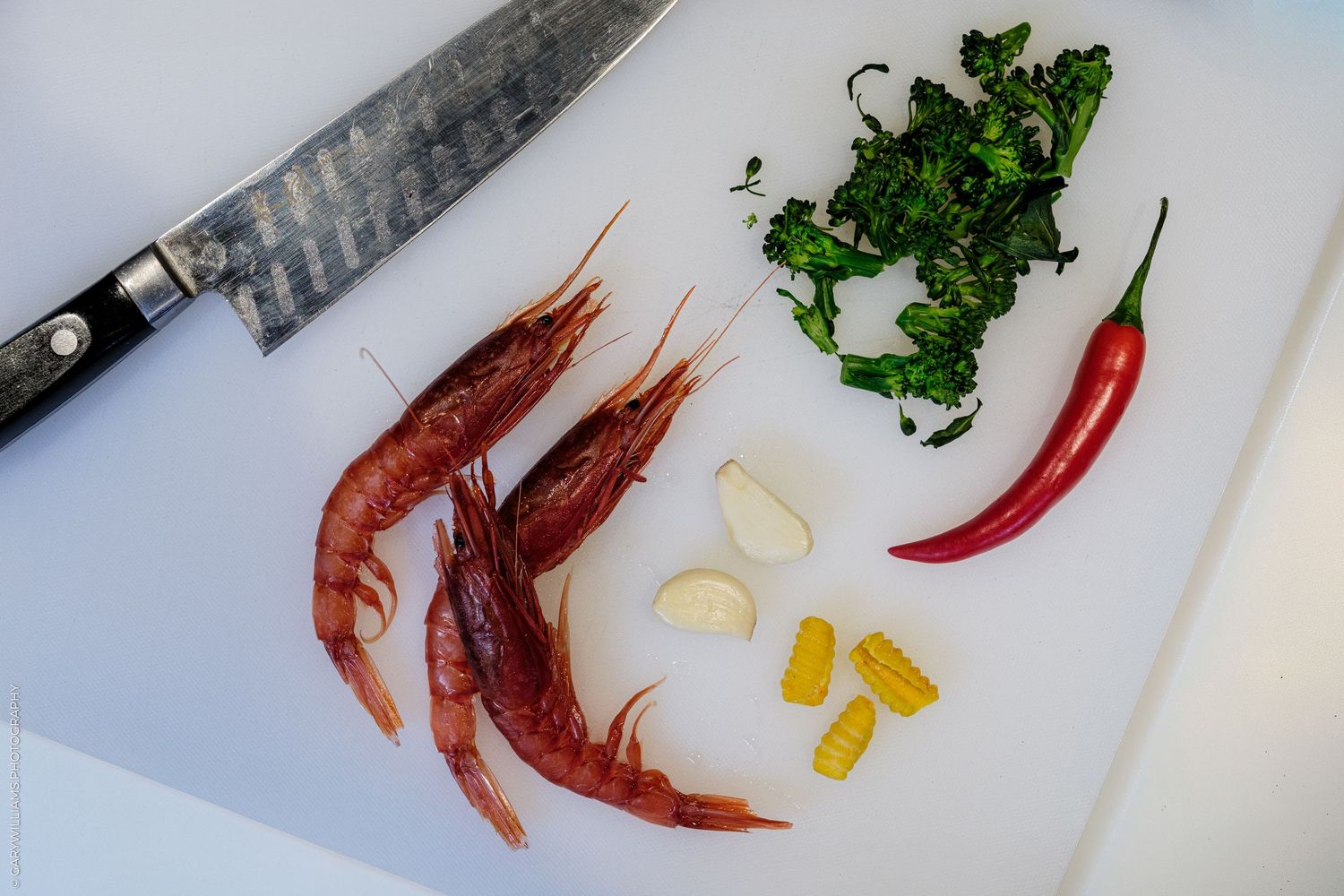 Raw shrimp, garlic, red chili, chopped herbs on white cutting board with chef knife, fresh seafood cooking ingredients.