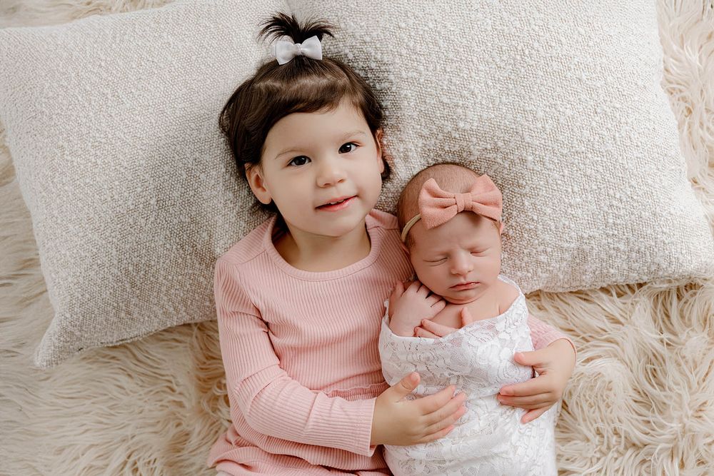 Young toddler girl in pink outfit holds sleeping newborn baby wrapped in white lace on a fluffy cream rug.