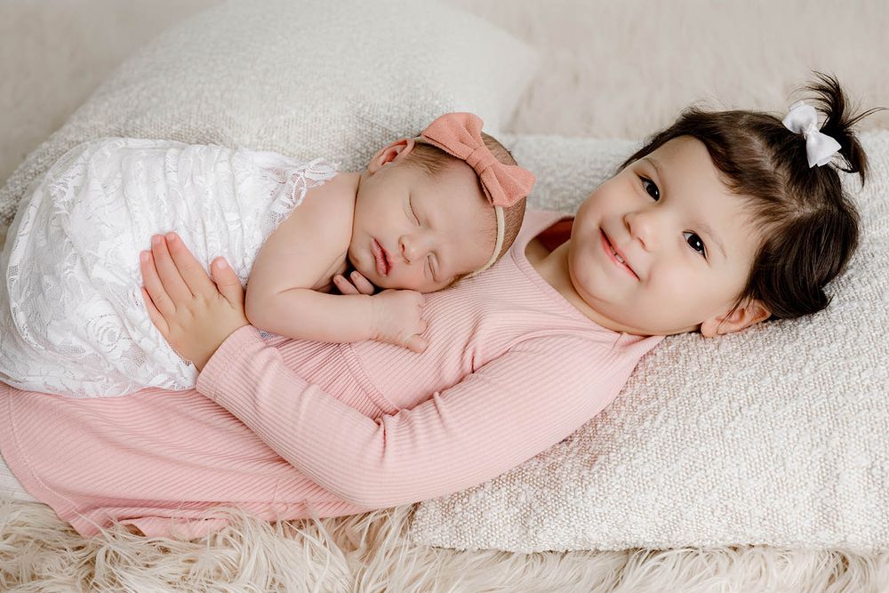 Two young sisters in pink outfits, older toddler holding sleeping newborn baby with bow headband on white fluffy blanket.