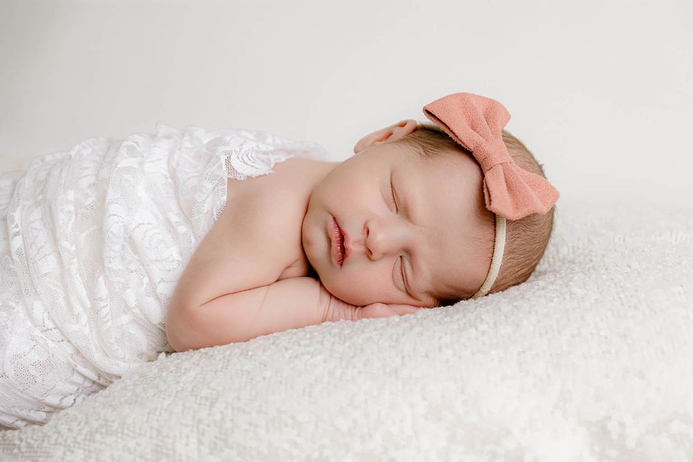 Peaceful newborn baby girl sleeping wrapped in white lace with a pink bow headband on a fluffy white blanket.