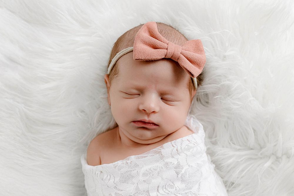 Sleeping newborn baby swaddled in white lace wrap with pink bow headband on fluffy white fur background.
