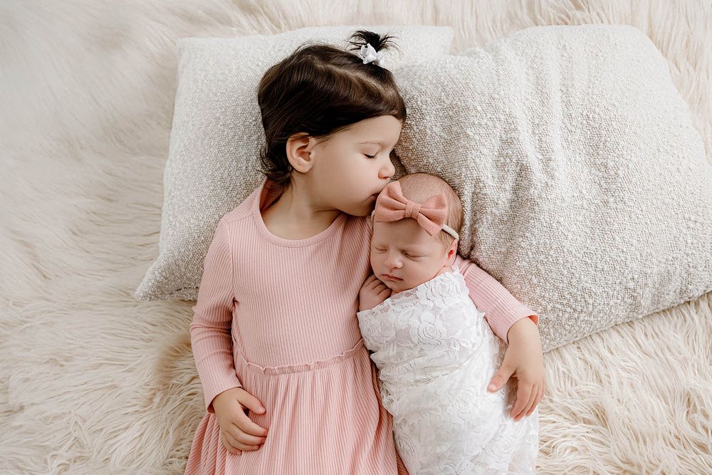 Older sister in pink dress kissing newborn baby sibling wrapped in white swaddle on fluffy cream rug.