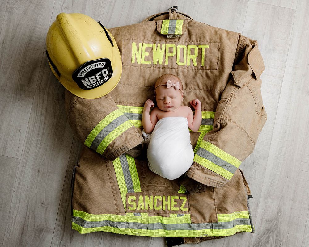 Newborn baby wrapped in white swaddle resting on firefighter turnout gear with Newport Sanchez name tags and yellow helmet.