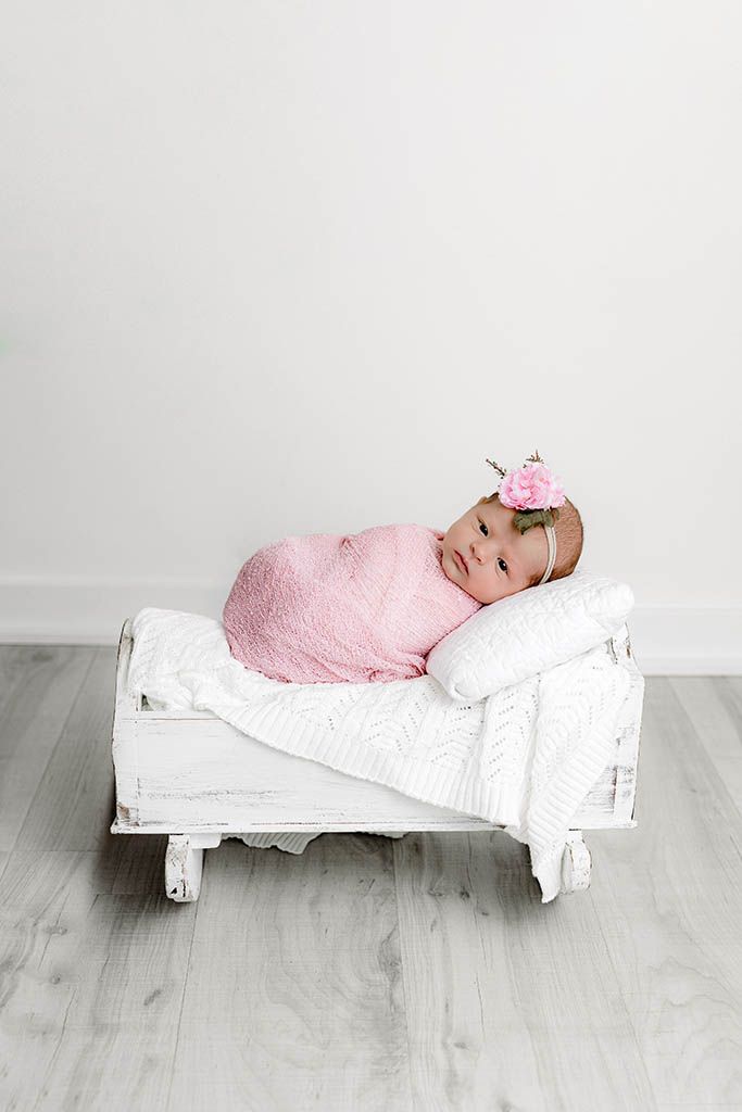 Newborn baby girl swaddled in pink wrap with floral headband, resting on a white miniature bed prop.