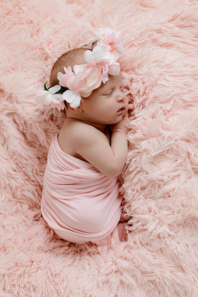 Newborn baby sleeping on pink fluffy blanket wrapped in pink swaddle with floral crown headband.