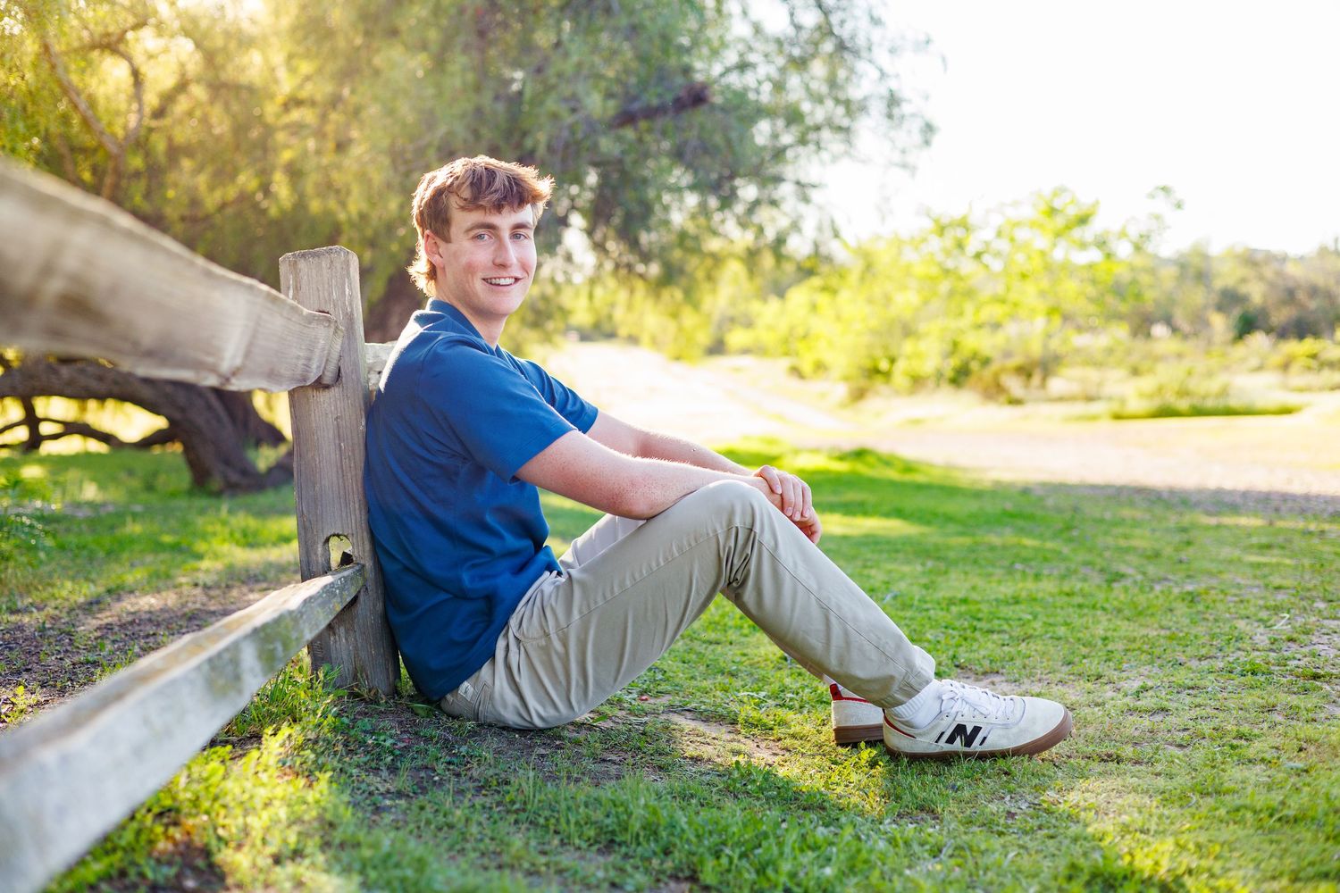 Young man in black polo shirt smiling with arms crossed outdoors on a sunny day.