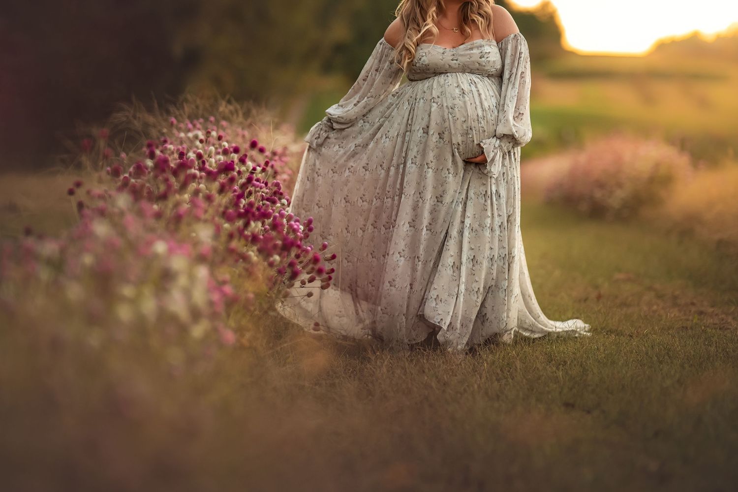 Pregnant woman in floral gown standing in a field of wildflowers during golden hour sunset.