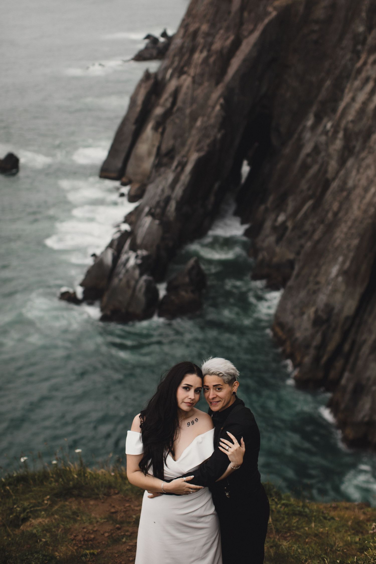 Dramatic coastal cliffs with green tops drop sharply into churning turquoise ocean waves below an overcast sky.