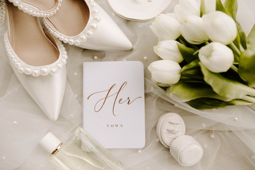 Elegant bridal flat lay with pearl heels, white tulips, perfume, ring box, and a gold script wedding vow booklet.