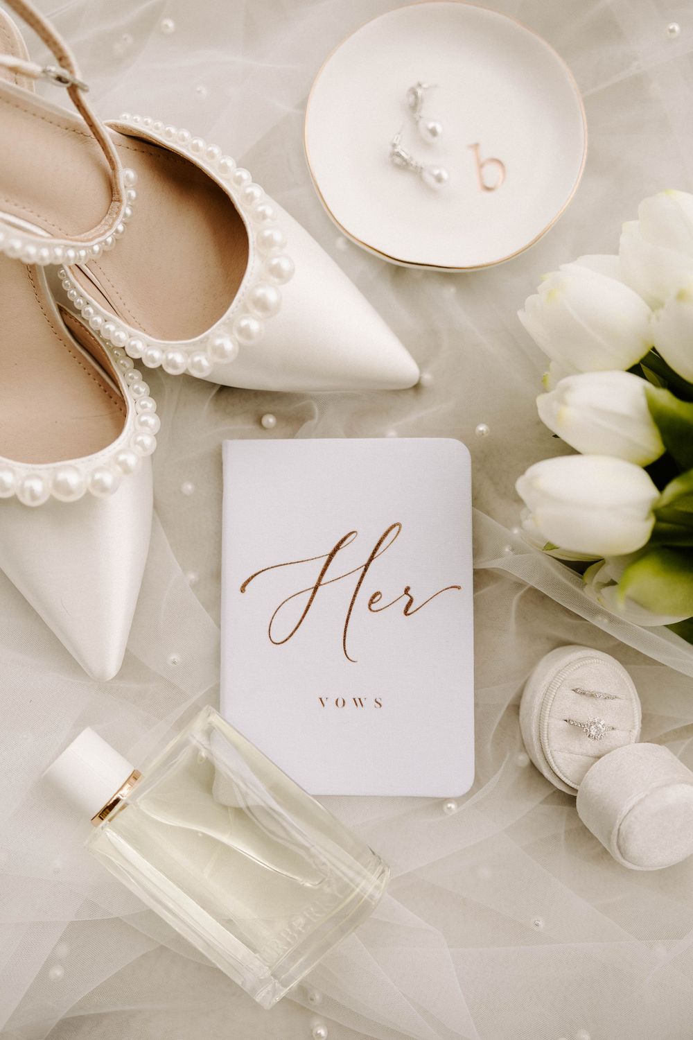 Elegant bridal flat lay featuring pearl heels, vow book, perfume, white tulips, and jewelry on soft white fabric.