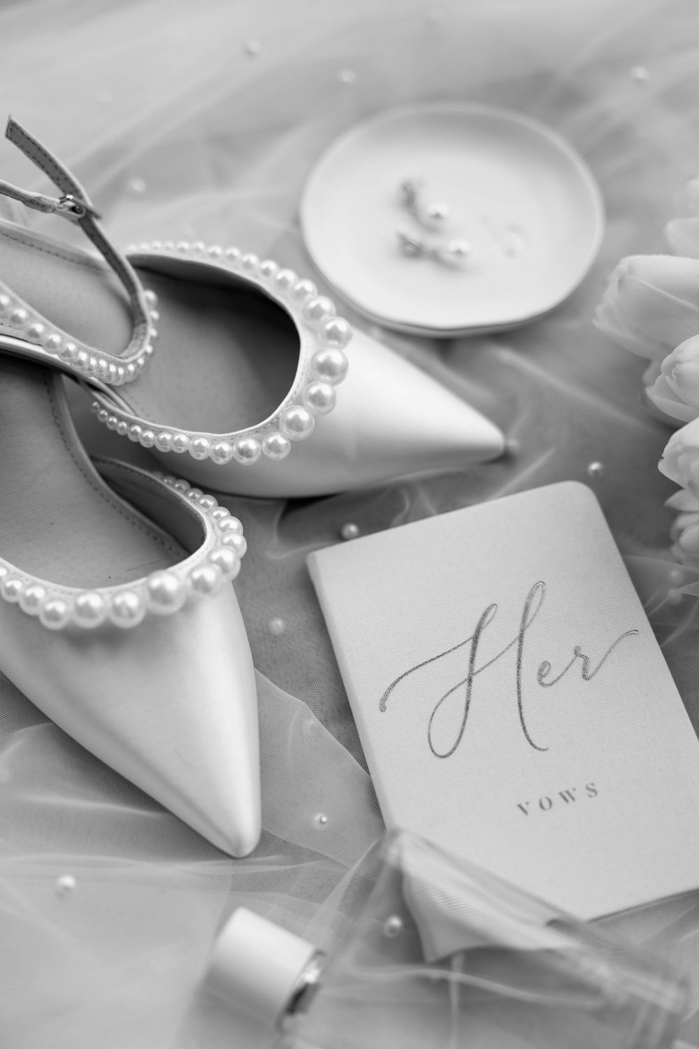 Black and white flat lay of pearl-trimmed bridal heels, her vows booklet, and delicate drop earrings on sheer veil.