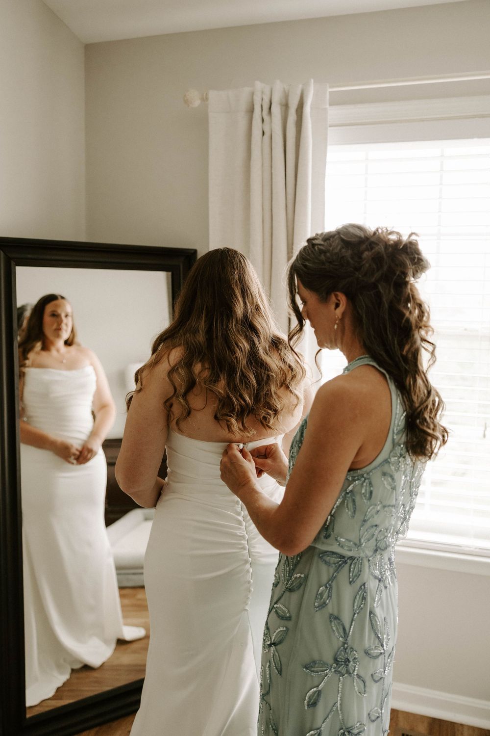 Bridesmaid helping bride button up her white wedding dress in front of a large mirror.