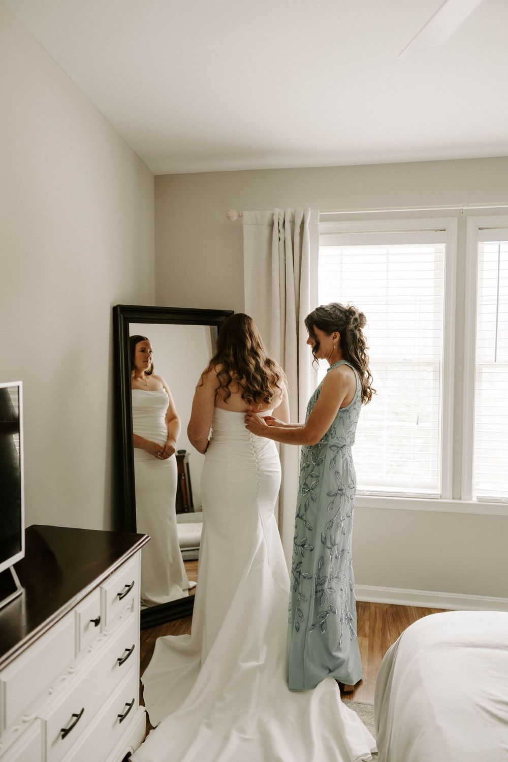 Bridesmaid in sage green dress helping bride into fitted white wedding gown in front of full-length mirror.