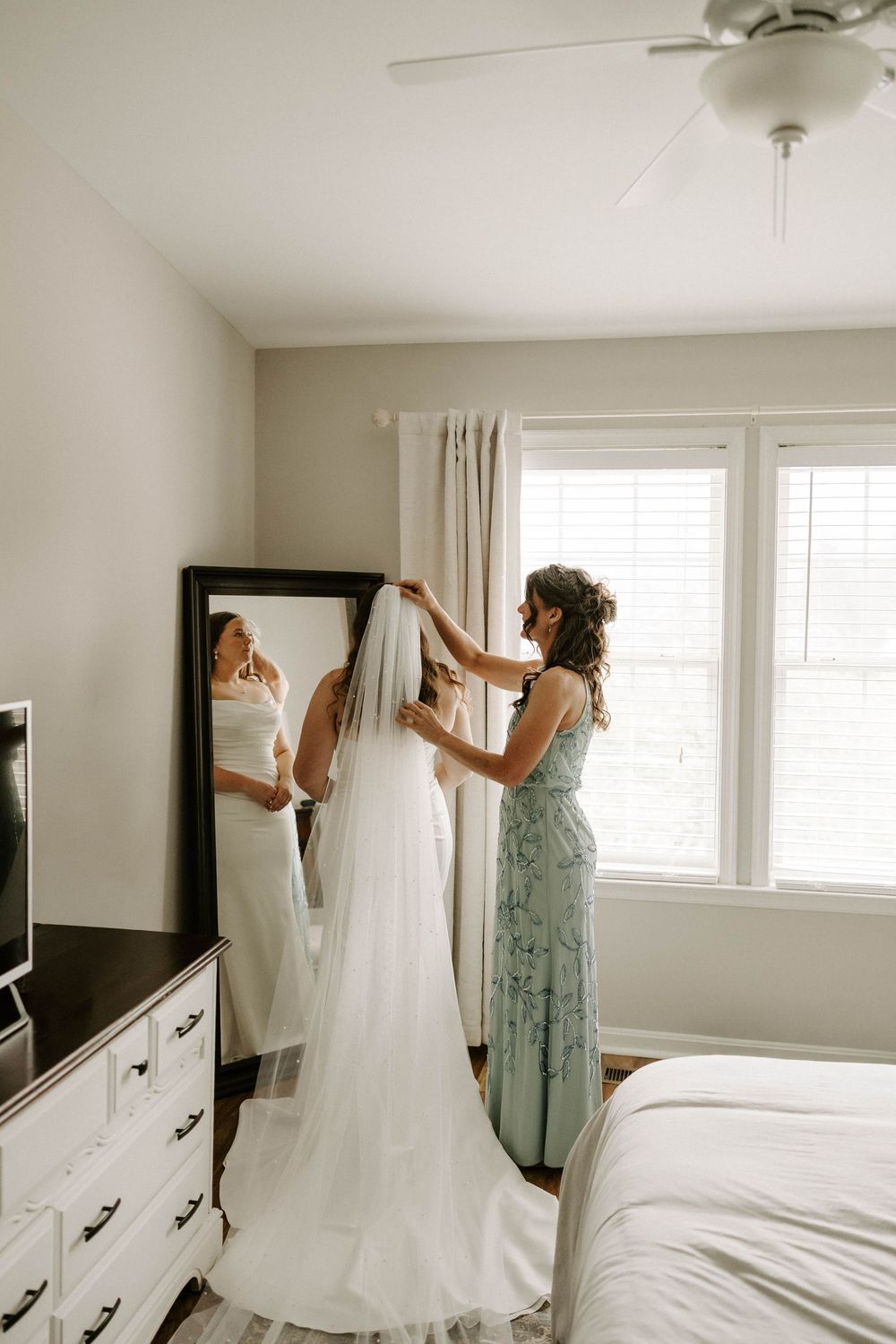 Bridesmaid helping bride with veil in bright bedroom before wedding ceremony.