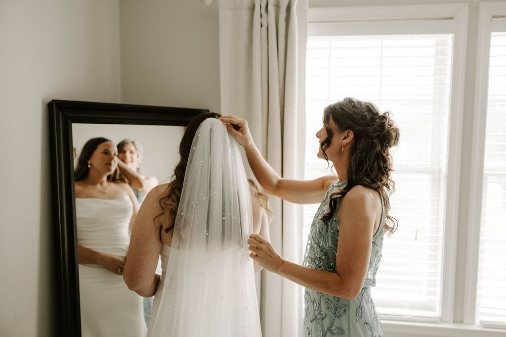 Bridesmaid helping bride adjust her veil in front of a mirror while getting ready for the wedding.