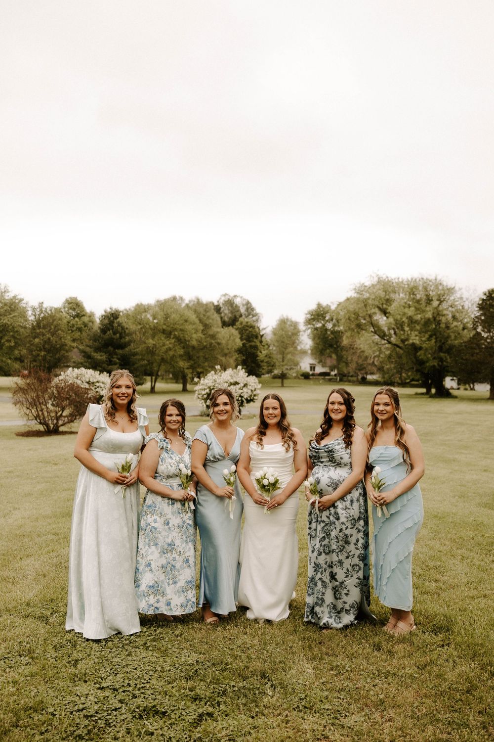 Bride with five bridesmaids in mismatched blue floral dresses holding bouquets on a green lawn.