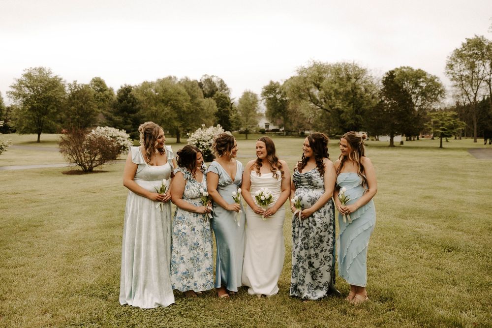 Bride in white gown poses with six bridesmaids in mismatched blue floral dresses on green lawn.