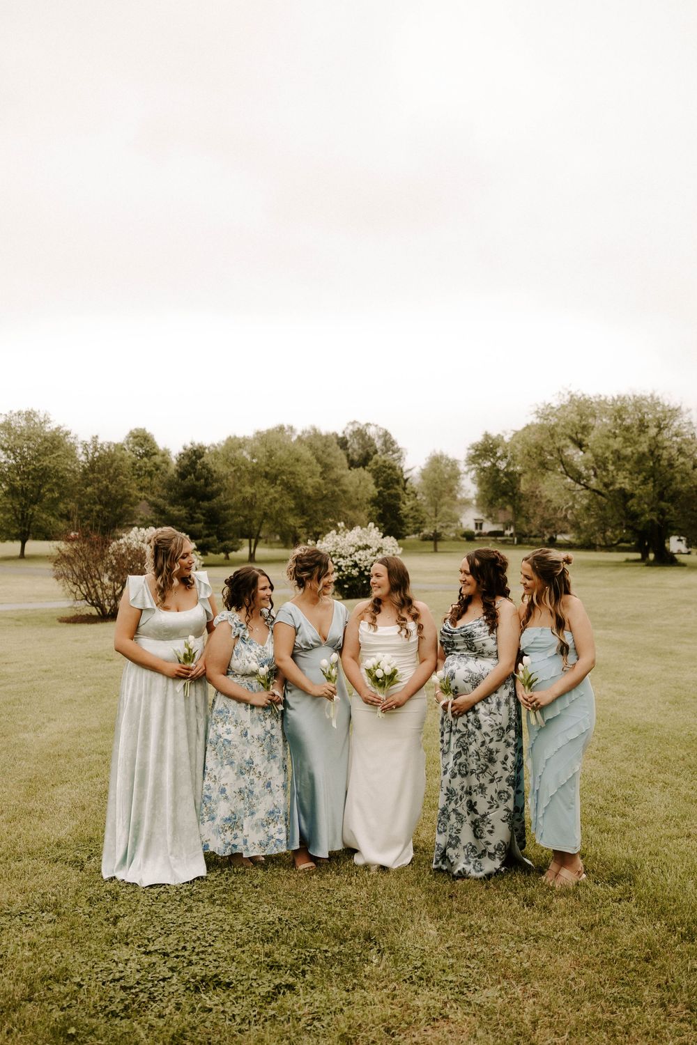 Bride in white gown surrounded by five bridesmaids in mismatched blue floral dresses on green lawn.