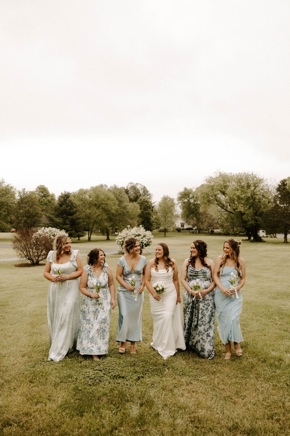 Bride and four bridesmaids in light blue and floral dresses walking outdoors on wedding day.