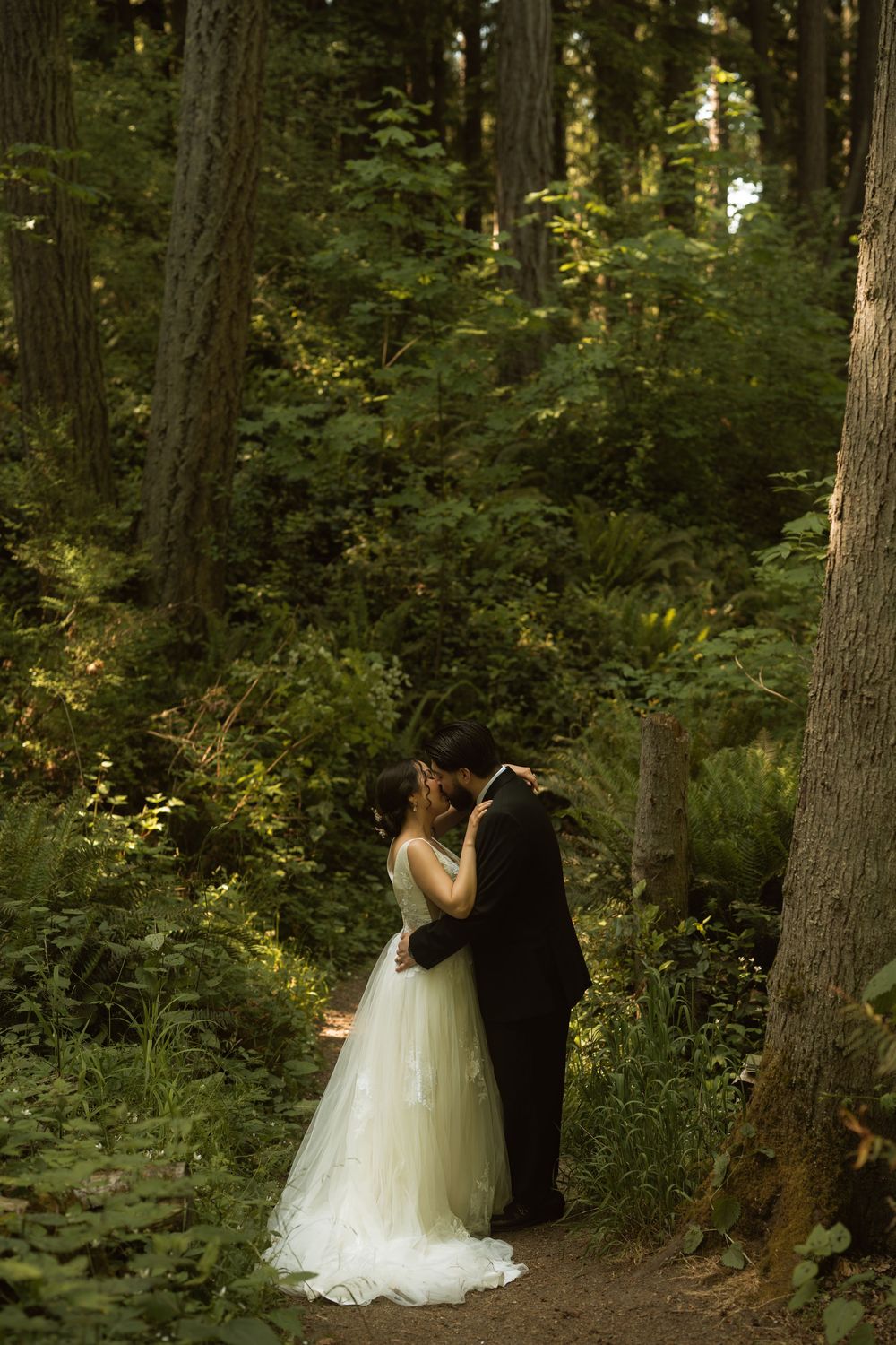 Bride and groom share a romantic kiss on a forest path surrounded by lush green trees and foliage.