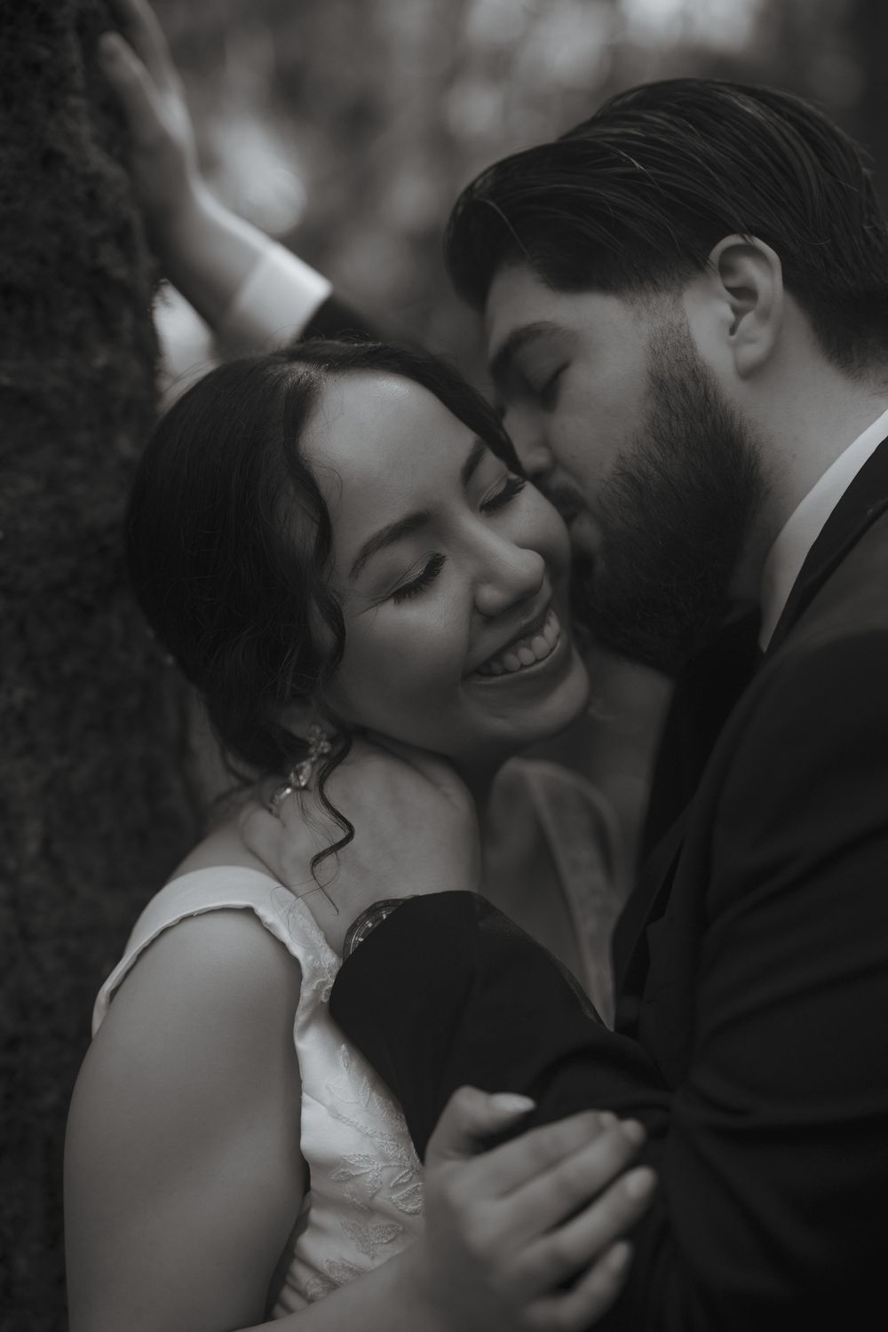 Black and white photo of a groom kissing his smiling bride on the cheek in a romantic forest wedding portrait.