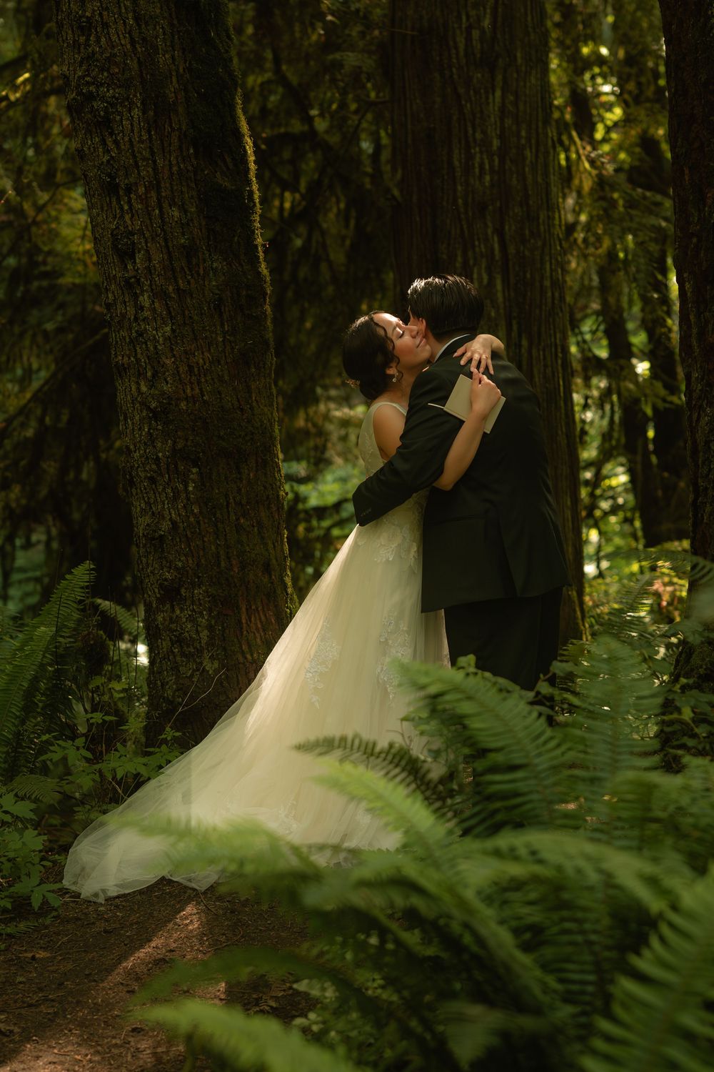 A bride and groom share a romantic kiss surrounded by towering trees and lush ferns in a forest setting.