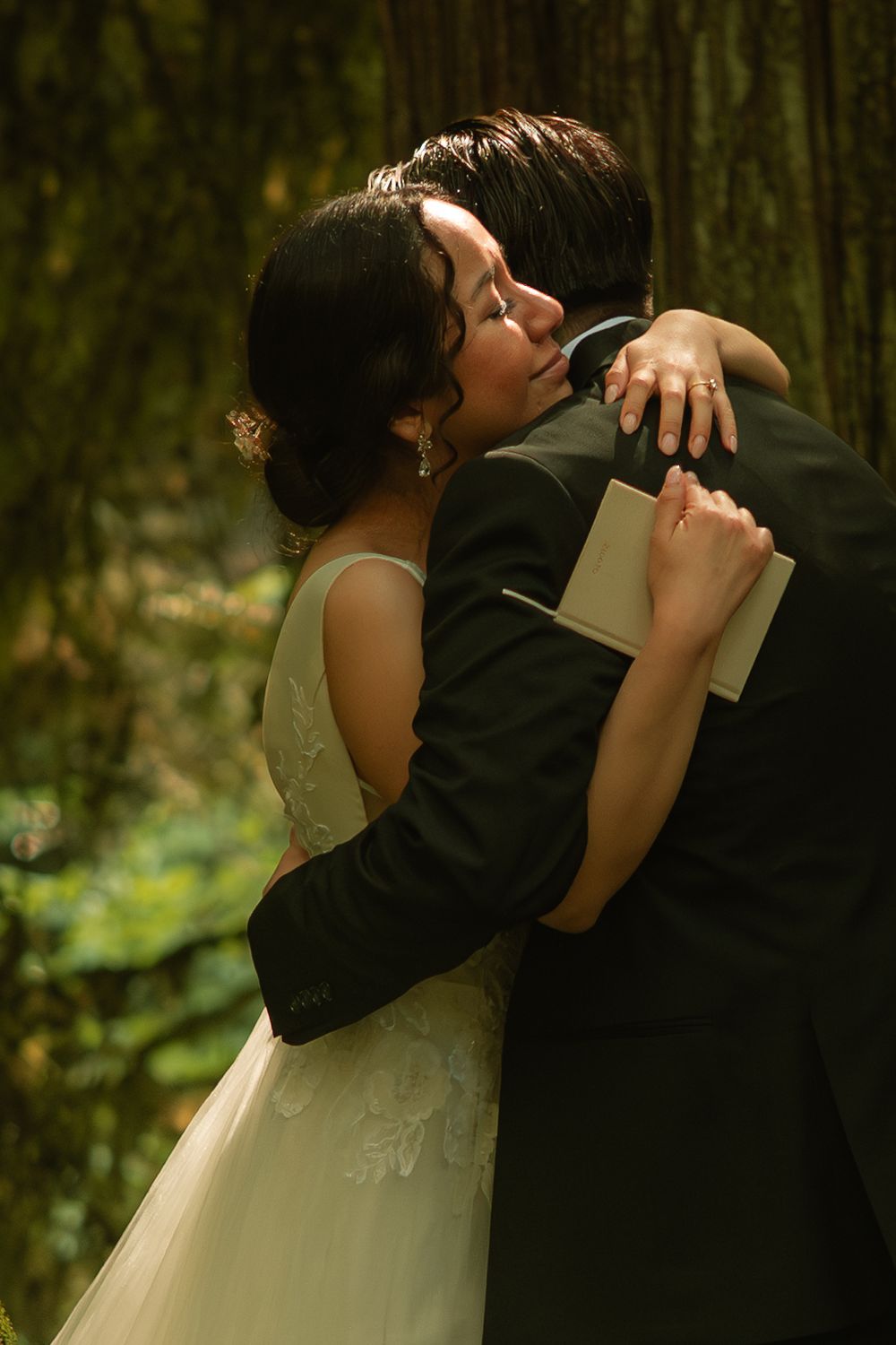 A bride and groom share a tender embrace in a lush forest setting, the bride wearing a white lace gown.