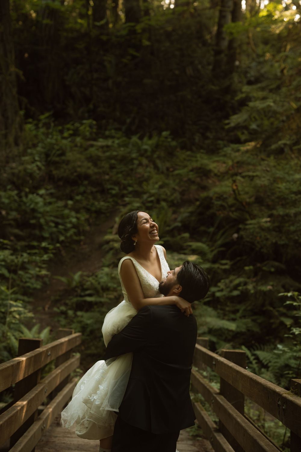 Groom lifting bride in white dress on forest bridge, both laughing joyfully among lush green trees.